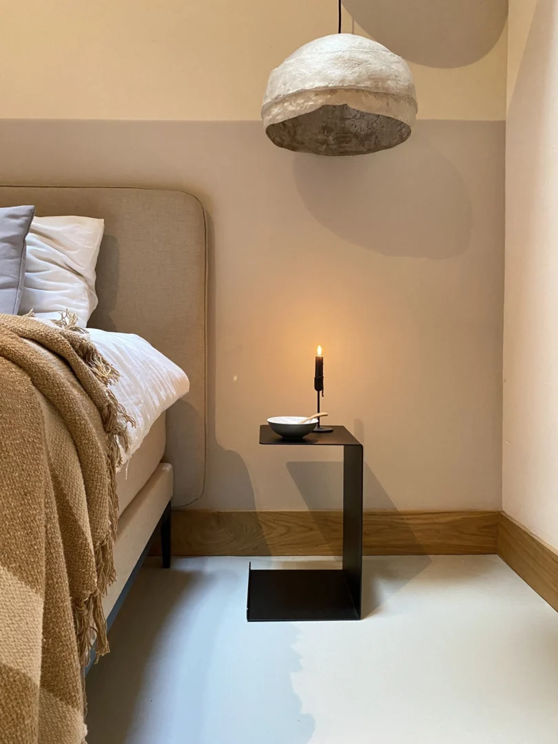 Een moderne slaapkamerhoek met een beige gestoffeerd bed, een geblokte bruine deken, een minimalistisch zwart bijzettafeltje met een brandende kaars en schaal en de koepelvormige hanglamp Meni30/20 - Standaard - Zwart die erboven hangt.