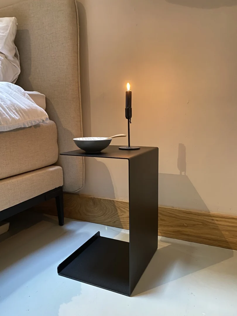De Meni30/20 - Standaard - Zwart, een moderne zwarte bijzettafel met een minimalistisch geometrisch ontwerp, staat op een lichte vloer bij een beige bed, met een brandende kaars in een houder en een witte schaal met lepel tegen een neutrale muur.