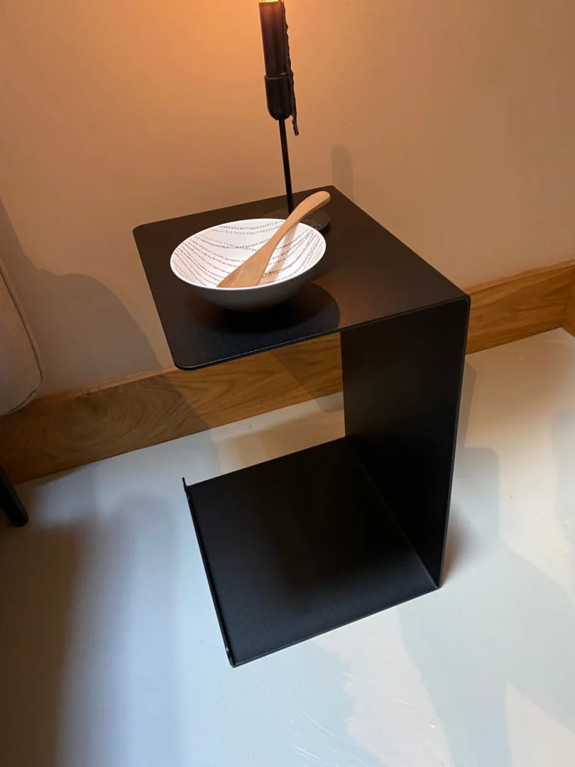 De Meni30/20 - Standaard - Zwart, een moderne zwart metalen C-vormige bijzettafel, houdt een witte kom en houten lepel onder een lamp op een lichtgekleurde vloer naast een beige muur met houten afwerking.