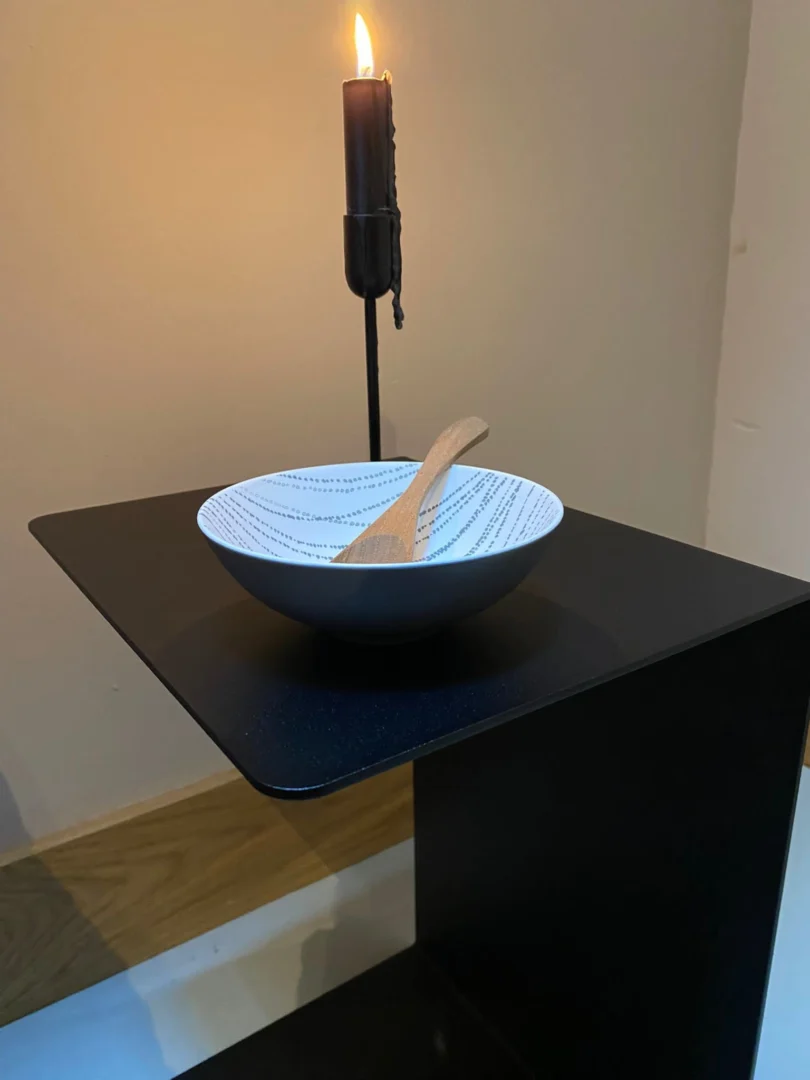 De kandelaar Meni30/20 - Standaard - Zwart met een brandende zwarte kaars staat achter een witte schaal met een patroon en een houten lepel, allemaal op een zwarte tafel tegen een effen muur.