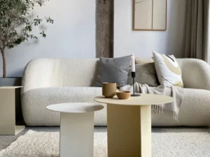 Een moderne woonkamer met een witte bank met structuur, neutrale kussens en een deken, twee Lucca Ronde bijzettafelsets in wit/crème, een potplant, abstracte muurkunst en een zachtbeige vloerkleed.