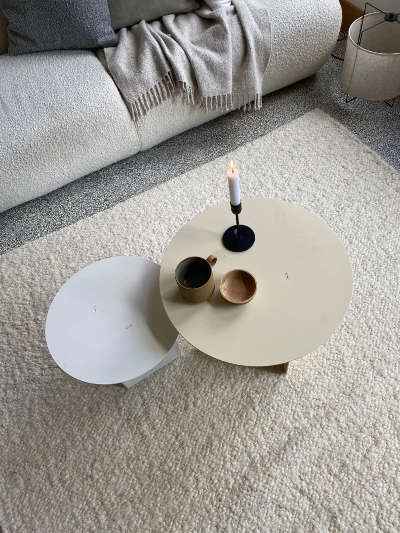 De Ronde bijzettafel set - Lucca - Wit/Crème bestaat uit een ronde beige salontafel met een zwarte kaarsenhouder, beker en houten schaal, gecombineerd met een kleinere witte bijzettafel op een gestructureerd crèmekleurig vloerkleed in een gezellige woonkamer.