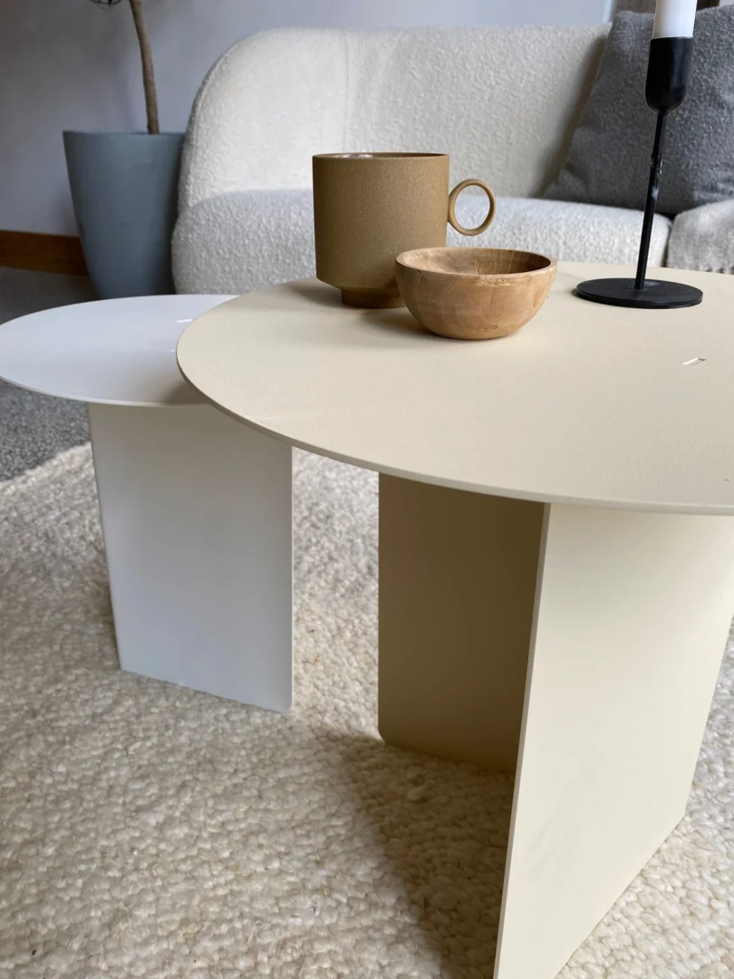 De Ronde bijzettafelset - Lucca - Wit/Crème bestaat uit twee minimalistische ronde tafels, een crème en een witte, op een beige vloerkleed met een bruin kopje, houten schaal en zwart-witte kandelaar. Op de achtergrond staan een bank en een plant.