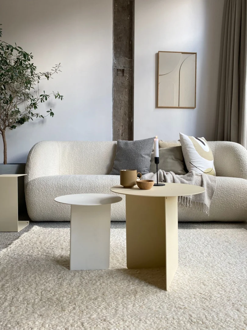 Een moderne woonkamer met een witte bank met textuur, neutrale kussens, twee Ronde bijzettafelset - Lucca - Wit/Crème, een potplant, een kaars, keramiek en abstracte muurkunst in zachte beige en grijze tinten.