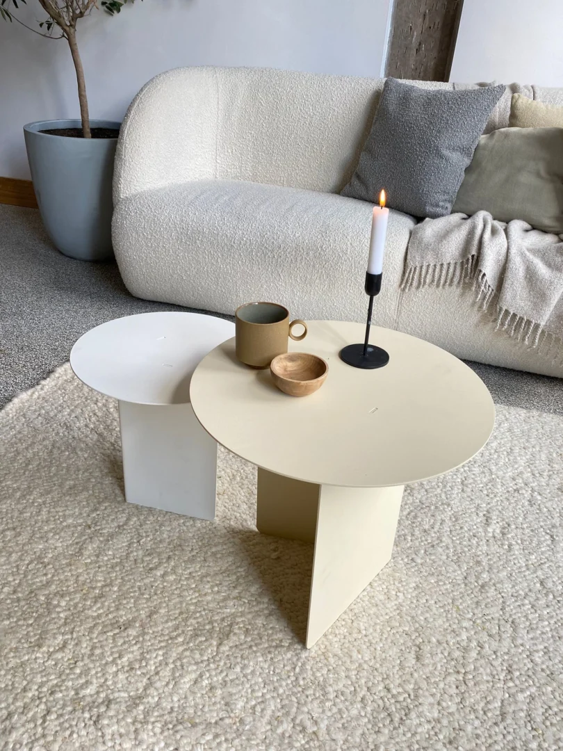 De Ronde bijzettafelset - Lucca - Wit/Crème, met twee moderne bijzettafels in wit en crème, staat op een vloerkleed met textuur, een beige mok, een houten schaal en een brandende kaars. Een gezellige bank met kussens verschijnt op de achtergrond.