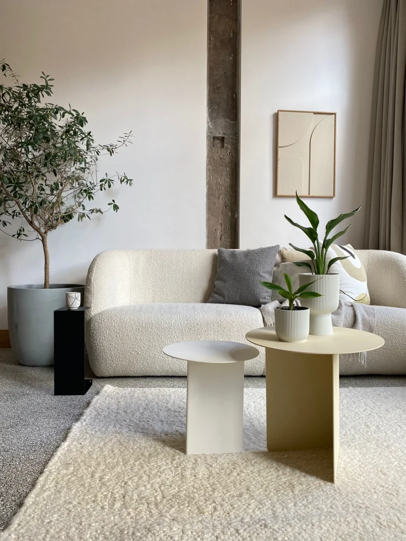 Een moderne woonkamer met een gestructureerde witte bank, neutrale kussens, twee Lucca Ronde bijzettafelsets in wit/crème met potplanten, een grote binnenboom, abstracte muurkunst en zacht natuurlijk licht vanuit een raam met grijze gordijnen.