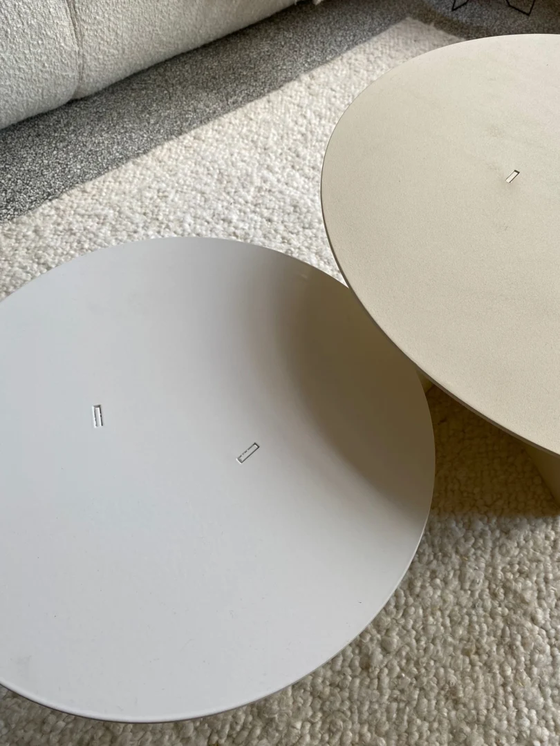 De Ronde bijzettafel set - Lucca in Wit/Crème heeft twee ronde, minimalistische salontafels op een gestructureerd gebroken wit tapijt, met kleine rechthoekige metalen accenten in het midden, naast een bank en grijze vloer op de achtergrond.