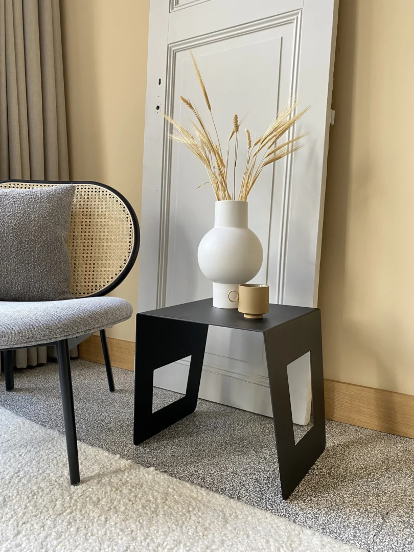 In een moderne woonkamerhoek staat de Brug - Bijzettafel - Zwart, gestileerd met een witte vaas met gedroogde tarwestengels en een beige kopje. Op de achtergrond staan een stoel met grijze structuur en een deur met decoratieve lijst.