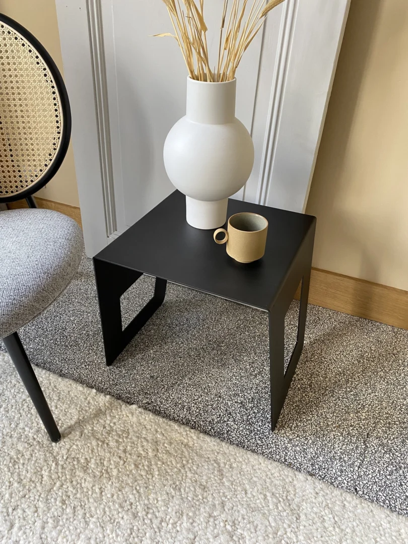 De Brug - Bijzettafel - Zwart heeft een matwitte vaas met gedroogd gras en een beige kopje erop. Hij staat op een tweekleurig vloerkleed met structuur naast een stoel met een geweven rieten rugleuning.