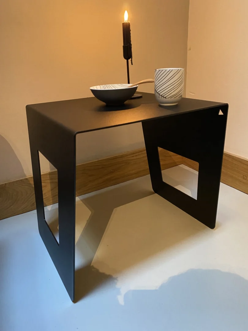 De Brug - Bijzettafel - Zwart, een compacte zwart metalen bijzettafel met geometrisch uitgesneden poten, toont een brandende zwarte kaars, witte kom en witte mok met zwarte diagonale lijnen tegen een lichte muur en houten plint.