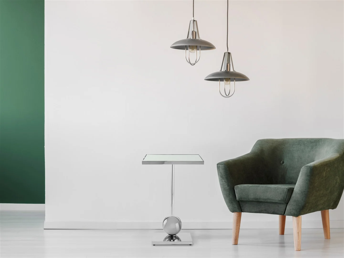 Een modern interieur met een groene fauteuil, de Bijzettafel Zilver Rond Staal Modern als strakke bijzettafel en twee hanglampen tegen een witte en groene accentmuur met lichte vloeren voor een minimalistische look.