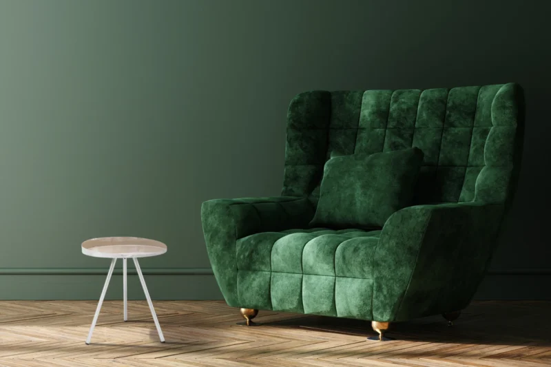 Een grote groene fluwelen fauteuil met een bijpassend kussen staat naast de Bijzettafel Taupe Ijzer Compact op een houten visgraatvloer tegen een donkergroene muur.