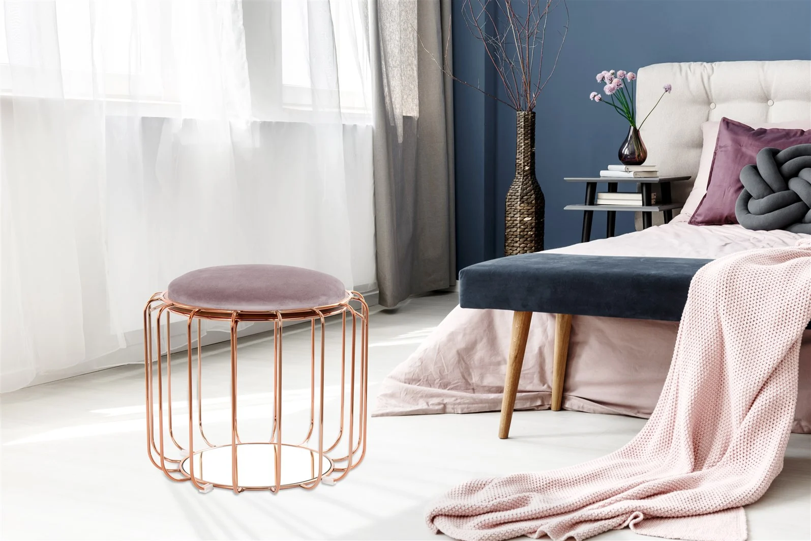Een moderne slaapkamer heeft een roze deken op het bed, een blauwe bank en een Bijzettafel Glas die wordt gebruikt als bijzettafel met boeken en een vaas, plus een rond metalen krukje met een roze kussen door witte gordijnen in het zonlicht.