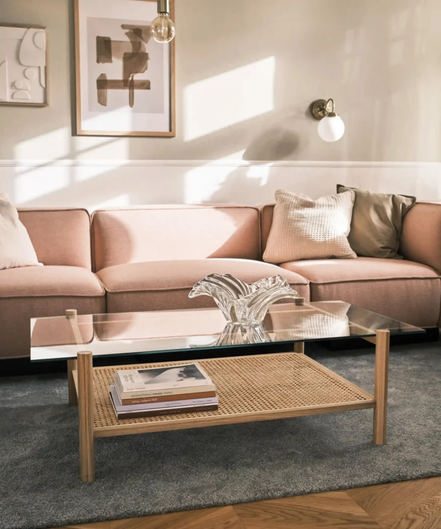 Een gezellige woonkamer met een lichtroze sofa, Salontafel 69 x 37 CM met boeken en een glassculptuur, neutrale kussens, ingelijste muurkunst en zacht natuurlijk licht dat naar binnen valt.