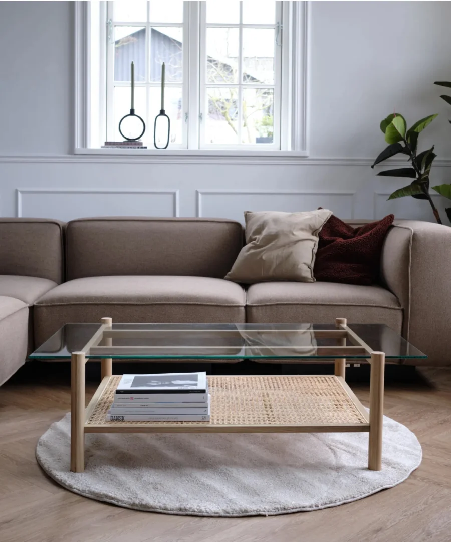 Een moderne woonkamer met een beige sectionele bank, Salontafel 69 x 37 CM, boeken, neutrale kussens, een rond vloerkleed, een potplant en kandelaars bij het raam creëren een gezellige minimalistische inrichting.
