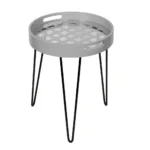 De Bijzettafel Grijs Metaal Compact is een kleine ronde bijzettafel met een lichtgrijs blad dat is voorzien van een geometrisch uitgesneden patroon en stevige zwarte metalen haarspeldpoten.