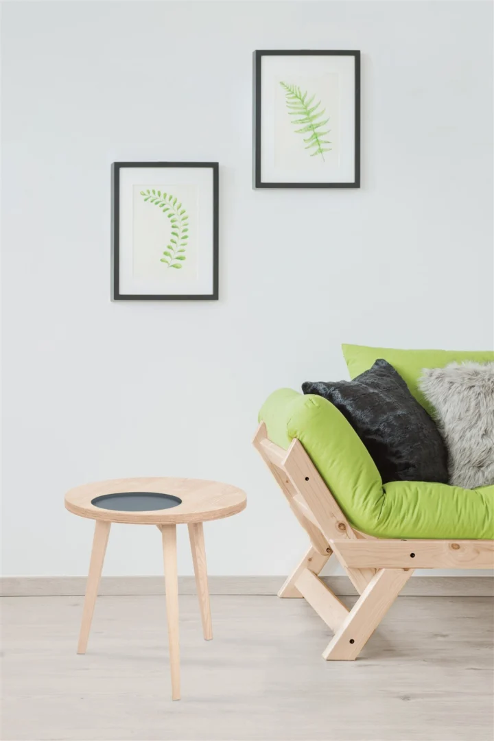 Een minimale woonkamer met een lichte houten vloer, een Bijzettafel Grijs Mdf als kleine ronde tafel, een houten bank met groene kussens en zwarte en grijze kussens, plus twee ingelijste varenprints op een witte muur.
