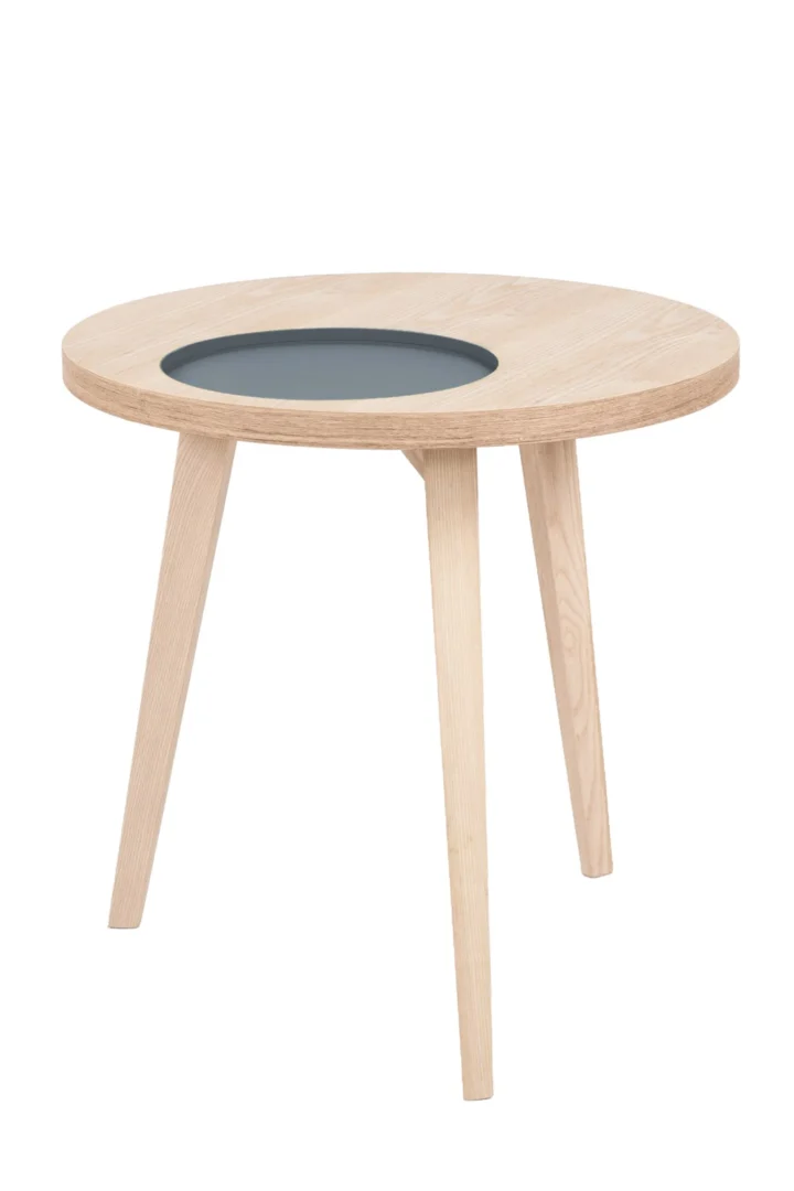 De Bijzettafel Grijs Mdf is een ronde, lichthouten bijzettafel met drie poten en een donkere ronde inleg in het midden van het tafelblad.