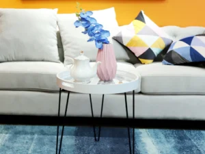 Een moderne woonkamer heeft een lichtgrijze bank, kussens met een patroon, de Bijzettafel Wit Metaal Design-een ronde witte salontafel met zwarte poten met daarop een roze vaas met blauwe bloemen, een witte theepot, een blauw vloerkleed en abstracte kunst op een oranje muur.