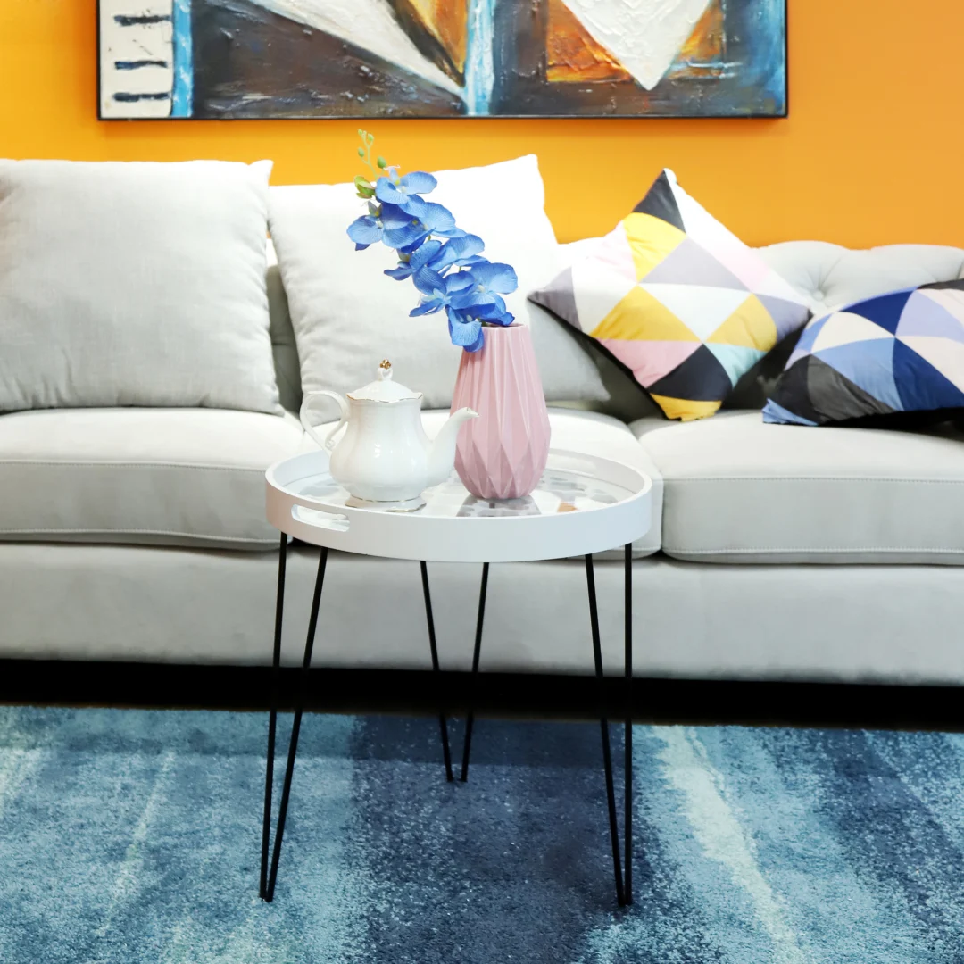 Een moderne woonkamer heeft een lichtgrijze bank, kussens met een patroon, de Bijzettafel Wit Metaal Design-een ronde witte salontafel met zwarte poten met daarop een roze vaas met blauwe bloemen, een witte theepot, een blauw vloerkleed en abstracte kunst op een oranje muur.