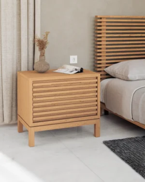 Het Nachtkastje Glas 41 x 61 x 53 CM staat naast een bed met een bijpassend hoofdeinde, met een vaas gedroogde bloemen en een tijdschrift. Op de vloer ligt een grijs vloerkleed, aangevuld met lichte gordijnen op de achtergrond.