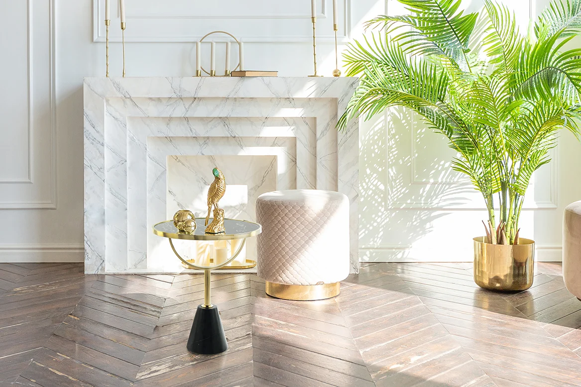 Lichte, elegante leefruimte met een wit marmeren open haard, een gewatteerde poef, een grote groene potplant op een visgraat houten vloer en de Bijzettafel Zwart, Roest Marmer. Zonlicht stroomt door een raam.