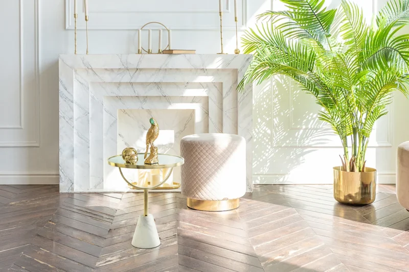 Een lichte, moderne woonkamer met een wit marmeren open haard, een gewatteerde kruk, een Bijzettafel Roest met wit marmeren blad en decor, en een grote palm in pot op een vloer van donker visgraathout.