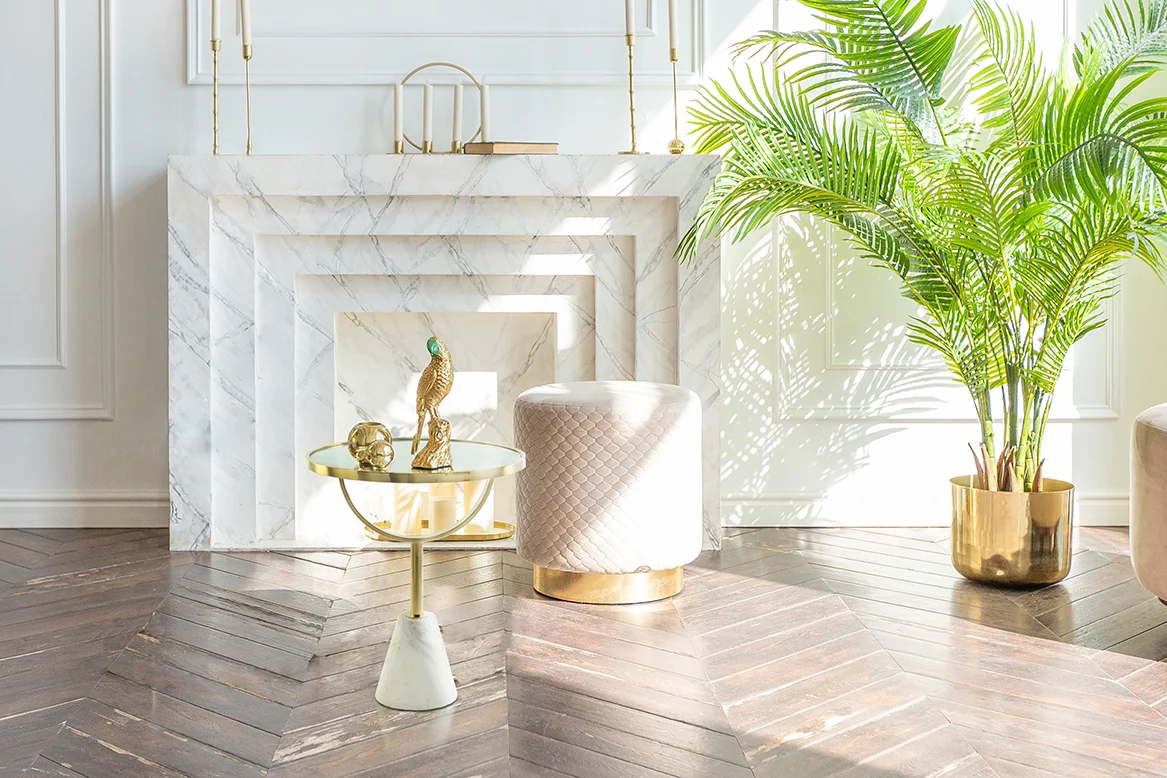 Een lichte, moderne woonkamer met een wit marmeren open haard, een gewatteerde kruk, een Bijzettafel Roest met wit marmeren blad en decor, en een grote palm in pot op een vloer van donker visgraathout.
