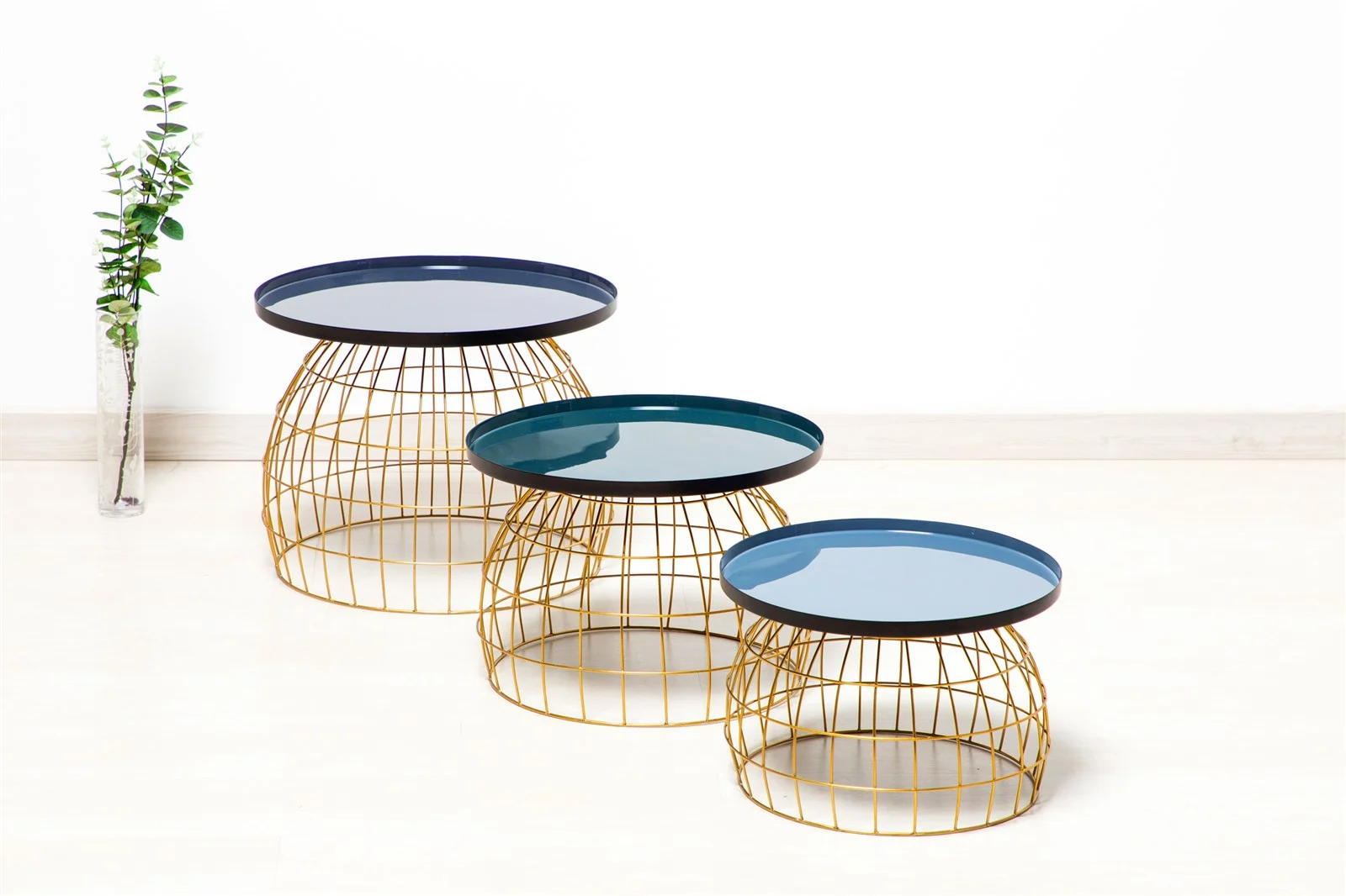 Drie Bijzettafel Grijs Rond tafels met gouden draadframe onderstellen en blauwe bladen zijn gerangschikt op hoogte van links naar rechts. Een kleine vaas met groen gebladerte staat links achter tegen een witte muur.