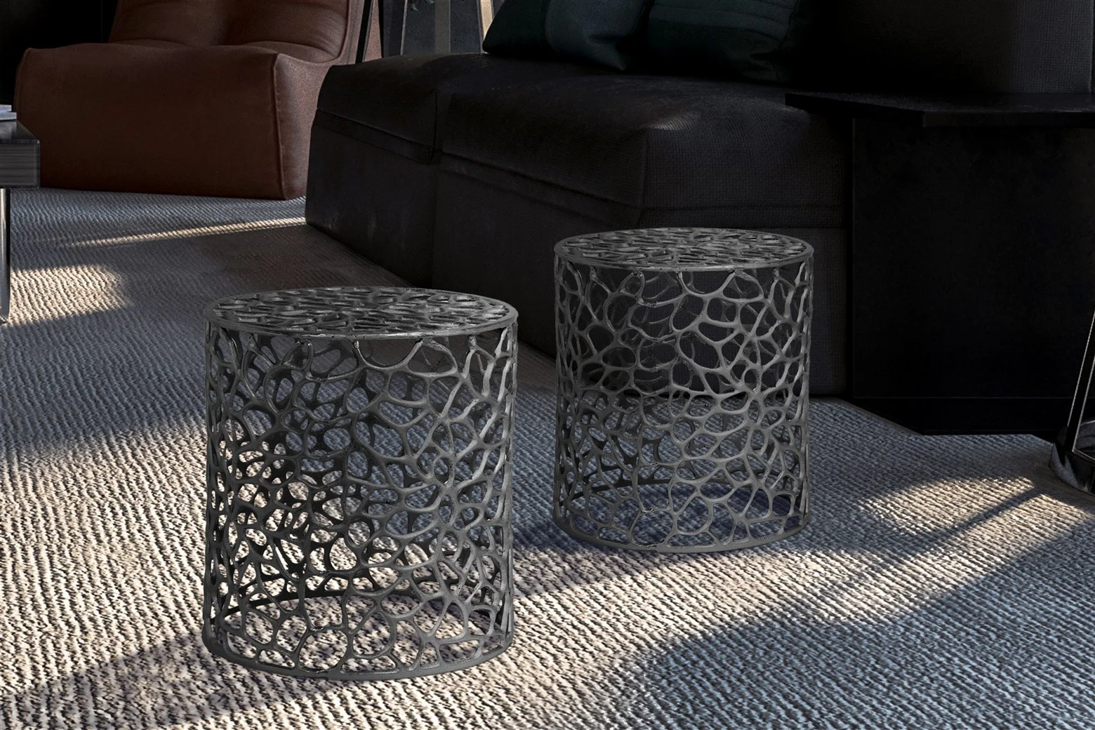 De Bijzettafel Zwart Aluminium Design, een ronde zwart metalen bijzettafel met opengewerkte patronen, staat op een lichtgrijs tapijt in een moderne woonkamer in de buurt van een donkere bank en een bruine fauteuil, waar het zonlicht schaduwen werpt op de vloer.