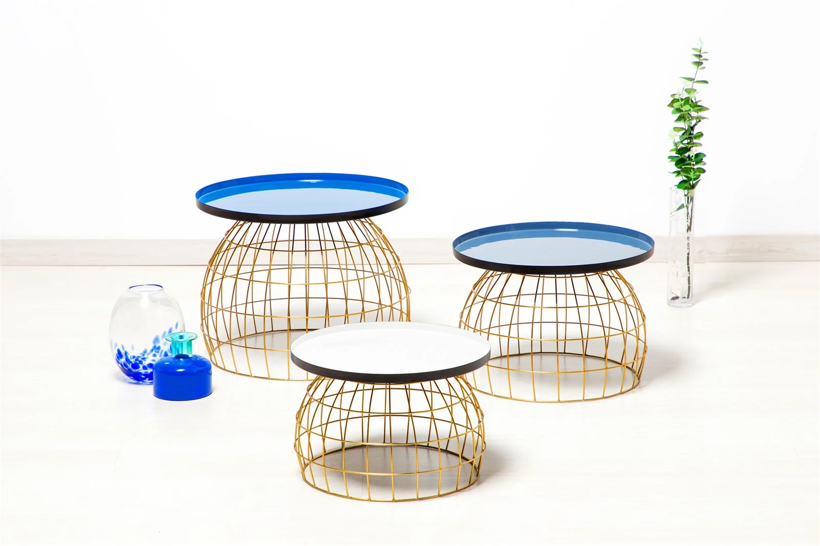 De Bijzettafel Grijs, Blauw bestaat uit drie ronde nesttafels met onderstellen van gouddraad en bladen van blauw en wit blad, gerangschikt op een lichte vloer naast twee decoratieve glazen vazen en een hoge vaas met groen gebladerte.