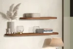 De SENTO BOEKENPLANK120 EL21 houten wandplanken tonen een vaas met gedroogd pampagras, een glas, gestapelde boeken en decor. Hieronder staat een beige tafellamp tegen een lichtgekleurde muur.