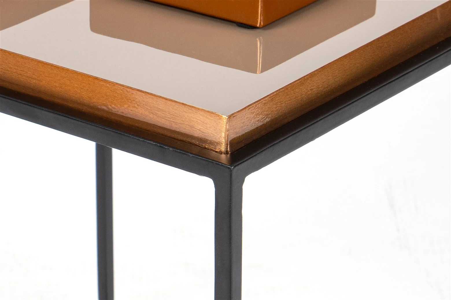 Close-up van de hoek van de Bijzettafel Crème Rond, met een glanzend lichtbruin houten blad met een reflecterend oppervlak en zwarte metalen poten. De rand van de tafel en het fijne vakmanschap zijn duidelijk zichtbaar.