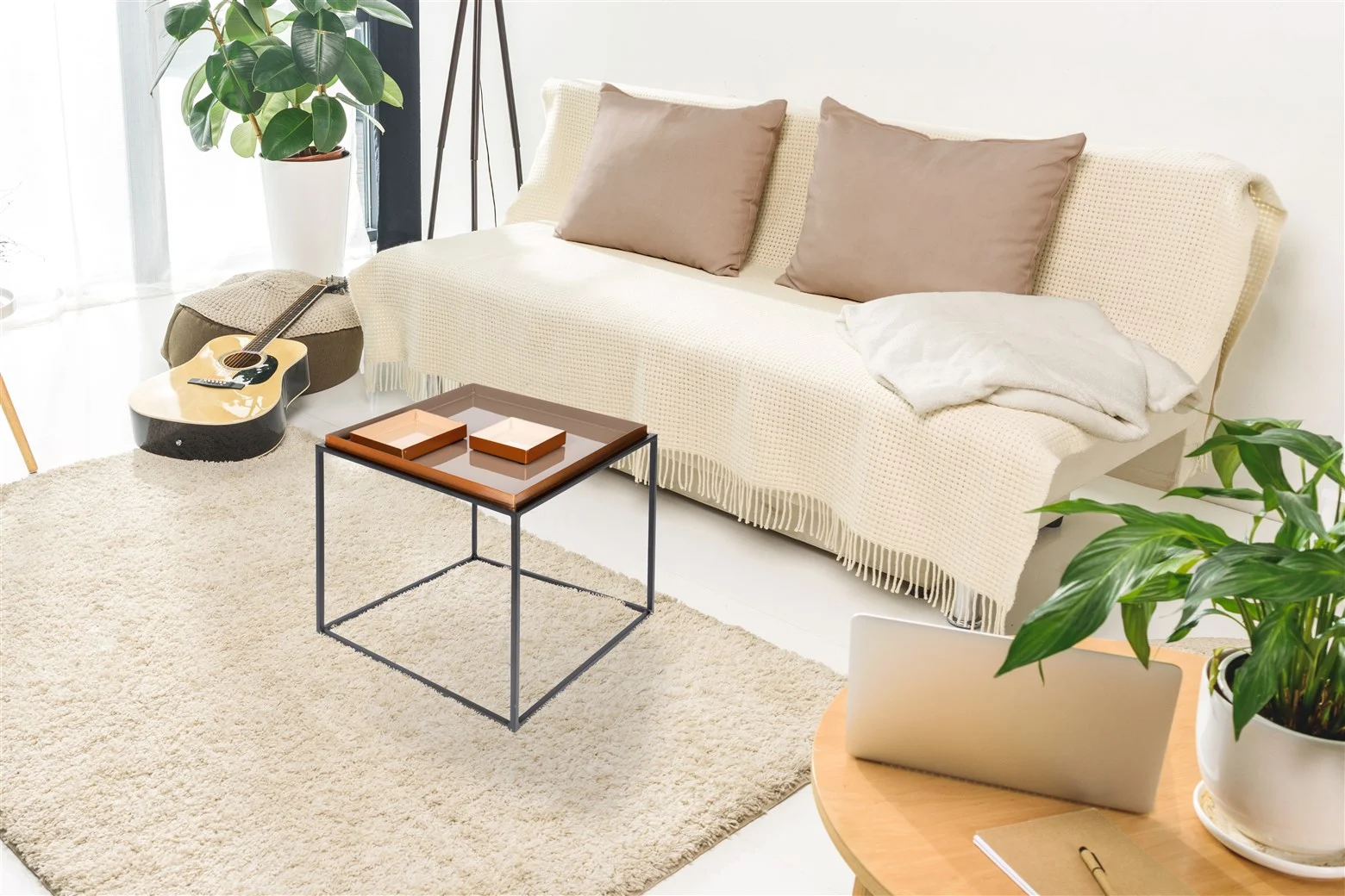 Een gezellige woonkamer met een witte bank, beige kussens en een deken. Een gitaar ligt in de buurt, groene planten sieren de kamer en een laptop staat op de Bijzettafel Crème Rond, waar ook een stapel boeken op ligt.