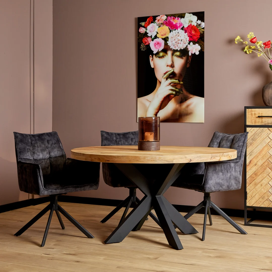 Een moderne eethoek met de Eettafel Denver, een ronde mangohouttafel (150 cm), drie zwarte draaistoelen, een middenstuk met kaarsen, muurkunst van een vrouw met bloemen op haar hoofd, roze muren en een houten vloer.