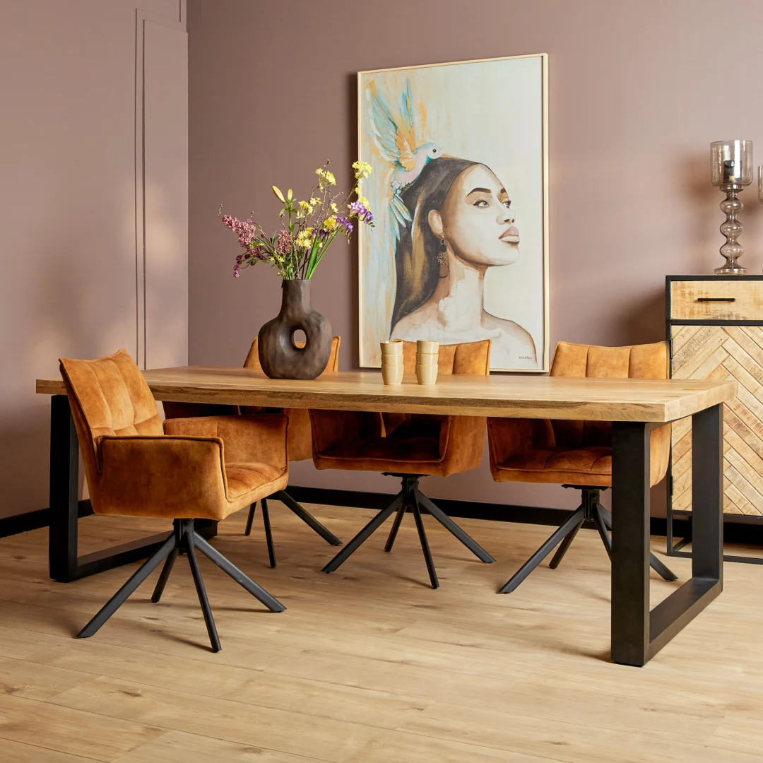 Een moderne eetkamer met de Eettafel Denver | Rechthoekige U-poot | 300 cm houten tafel, vier mosterdfluwelen stoelen met zwart metalen poten, een donkere vaas met bloemen, drie kopjes en een groot portret van een vrouw met een gevederde hoofdtooi op een beige muur.