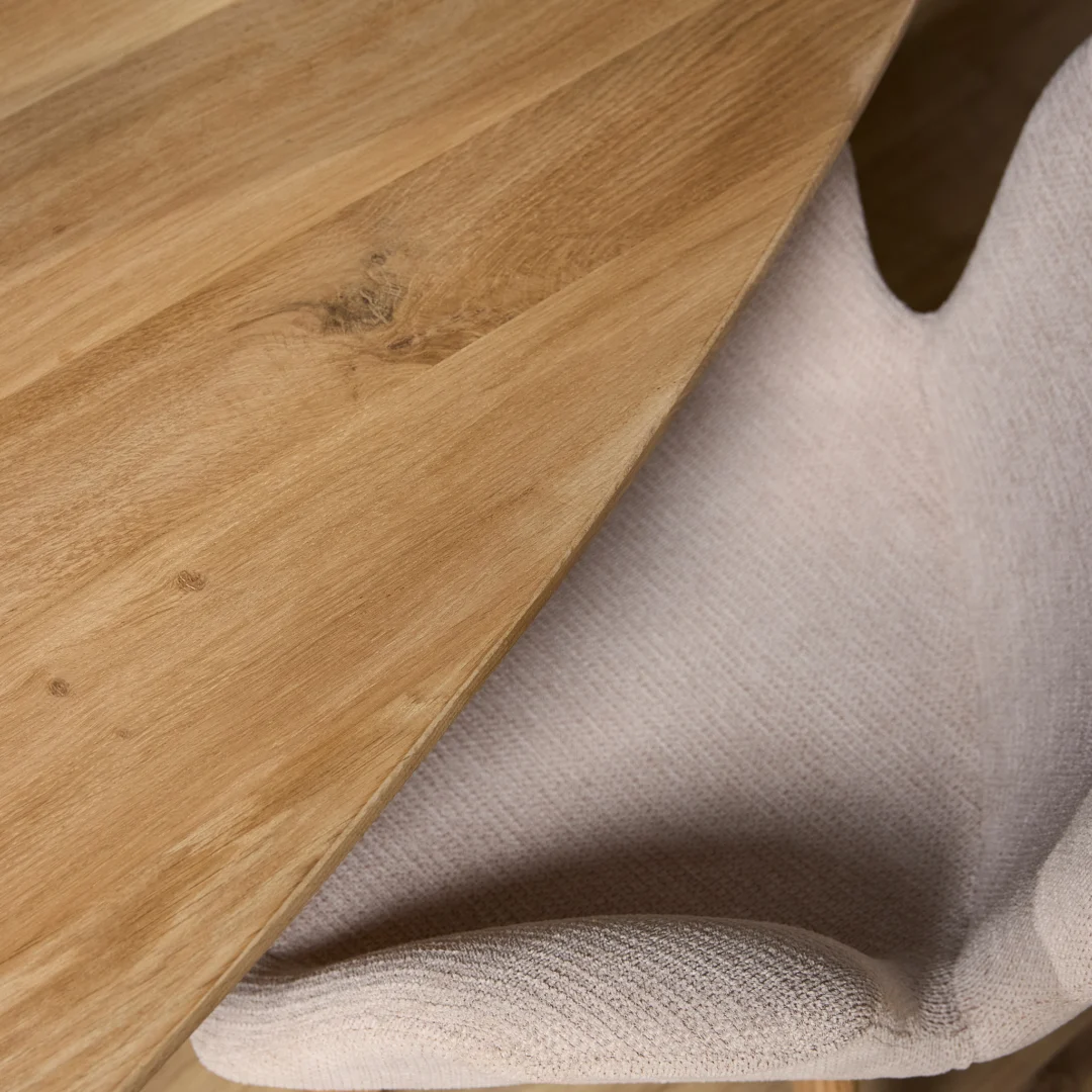 Close-up van een eikenhouten eettafelblad in naturel tinten, naast een zachte, witte stoel onder zacht licht.