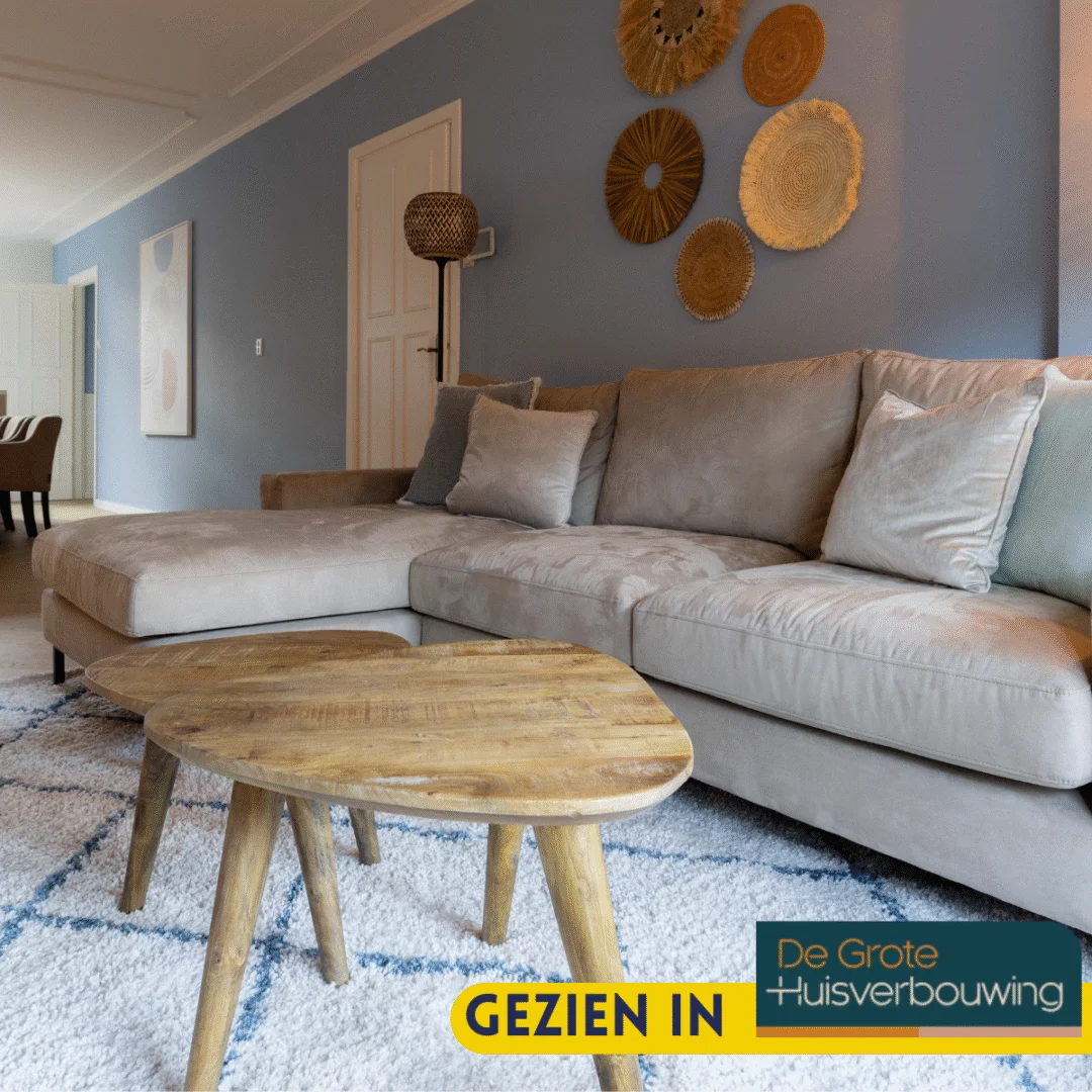 Een moderne woonkamer met een beige sectionele bank, lichtblauwe en grijze kussens, een Salontafel Design ronde houten salontafel, een gedessineerd vloerkleed, geweven wanddecoratie en lichtblauwe muren. Het logo van het tv-programma en de tekst GEZIEN IN verschijnen onderaan.