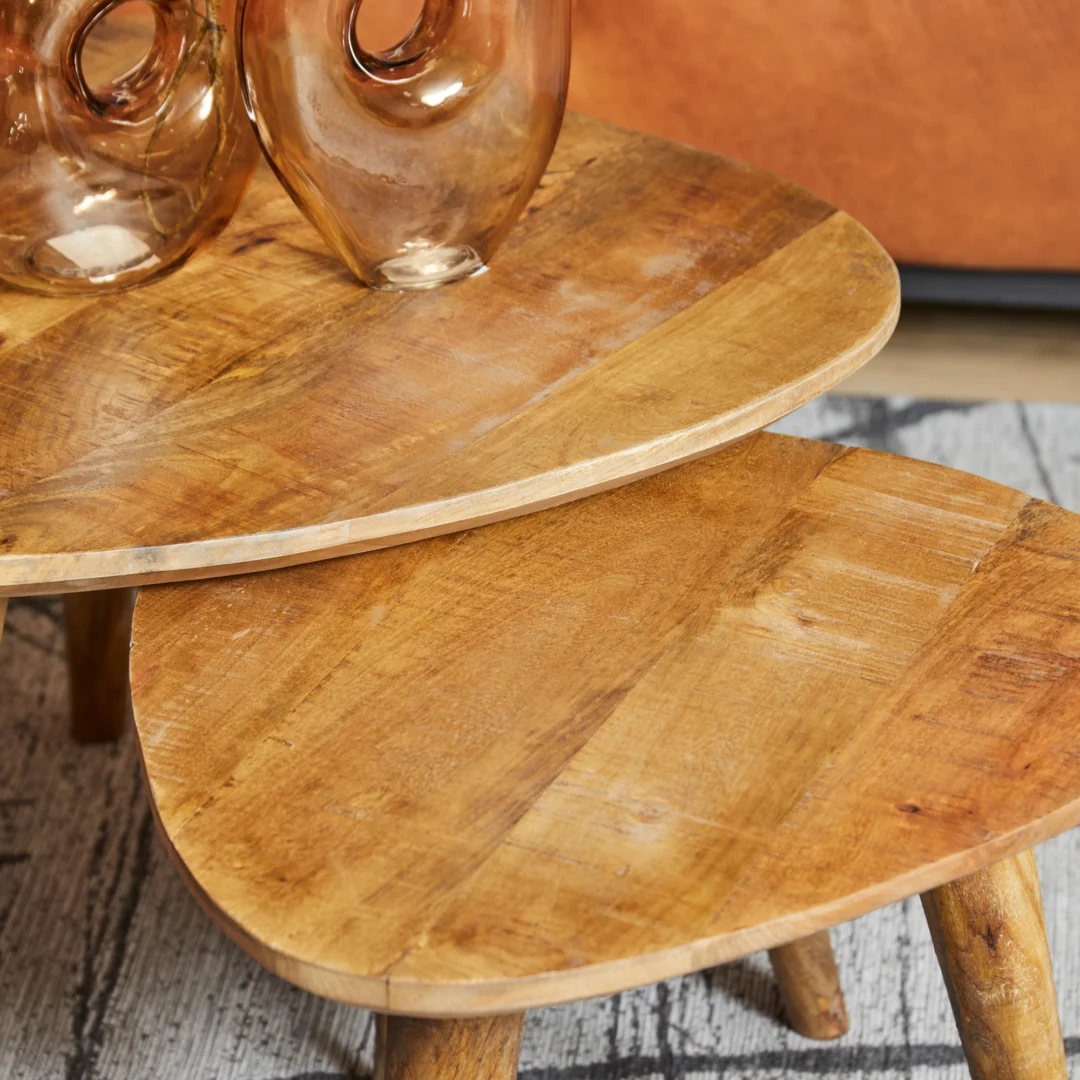 Twee overlappende Salontafel Design salontafels met afgeronde driehoekige houten bladen en zichtbare houtnerf. Glazen vazen staan op de bovenste tafel, die op een gedessineerd tapijt staat bij een bruine bank.