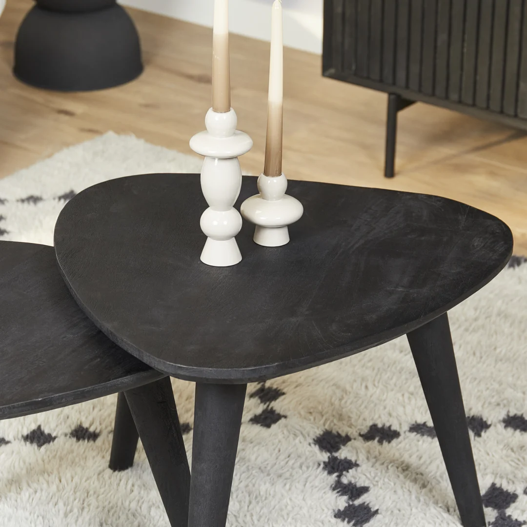 Een Vierkant Salontafel Zwart 66 x 56 CM houdt twee witte kandelaars met beige kaarsen vast. De vierkante zwarte salontafel staat op een tapijt met patroon, met een houten vloer en een zwarte kast op de achtergrond.