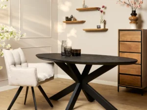 Een moderne eethoek met de Eettafel Brandy Zwart 160 cm ovale tafel en beige stoel met kussens. Houten planken met decor en een ladekast staan tegen een lichte muur, terwijl roze bloemen in een vaas voor een decoratieve touch zorgen.