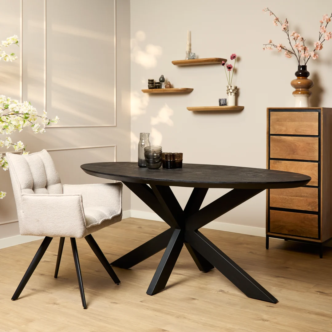 Een moderne eethoek met de Eettafel Brandy Zwart 160 cm ovale tafel en beige stoel met kussens. Houten planken met decor en een ladekast staan tegen een lichte muur, terwijl roze bloemen in een vaas voor een decoratieve touch zorgen.