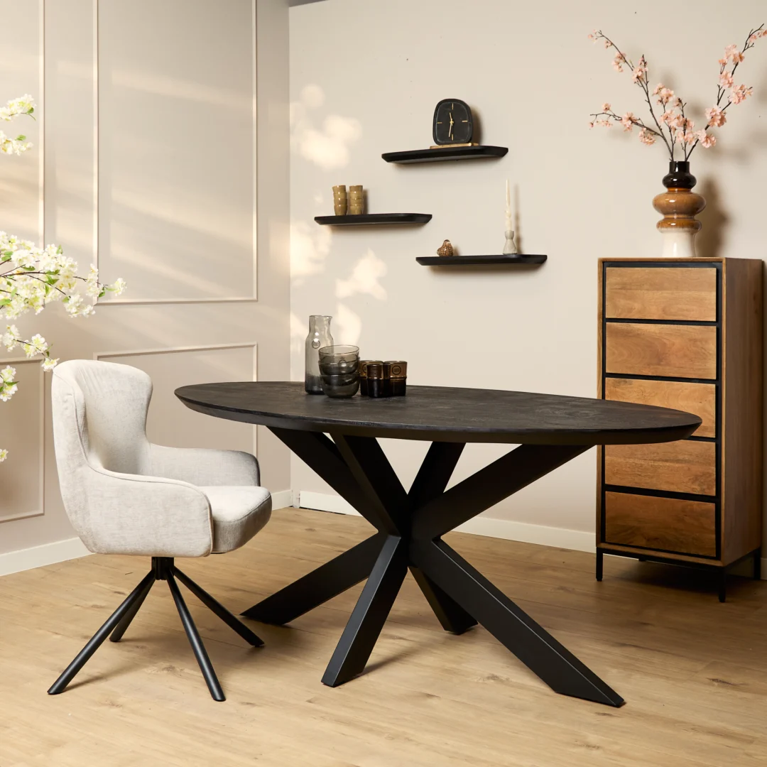 Moderne eetkamer met de Eettafel Brandy Zwart 160 cm ovale tafel, een beige draaistoel, houten dressoir, wandplanken met decor, lichthouten vloer en decoratieve takken en bloemen.