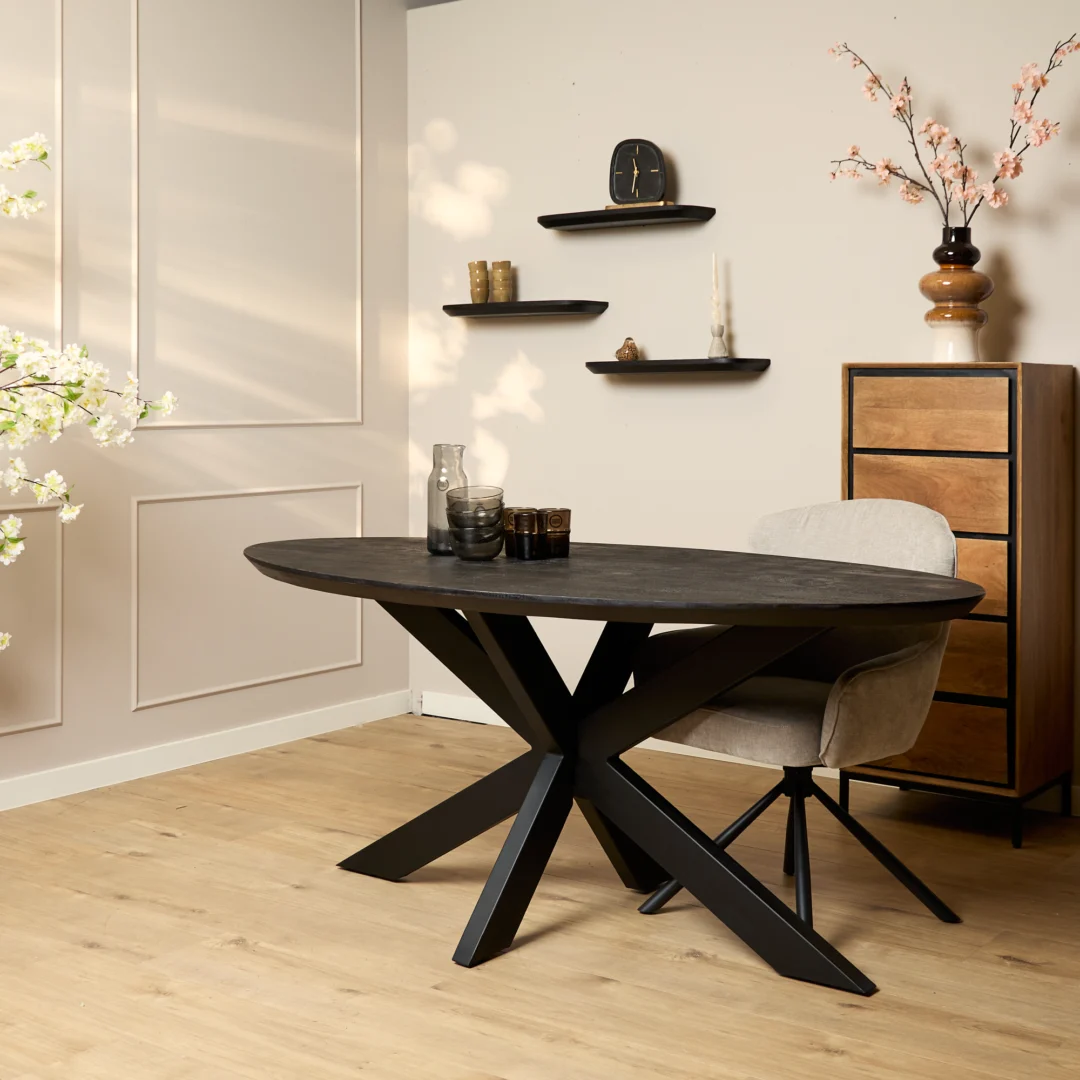 Moderne eetkamer met de Eettafel Brandy Zwart (160 cm, ovaal), beige gestoffeerde stoel, houten ladekast, zwevende planken met decor, bloesemtakken in een vaas, lichte houten vloer en warme crèmekleurige muren.