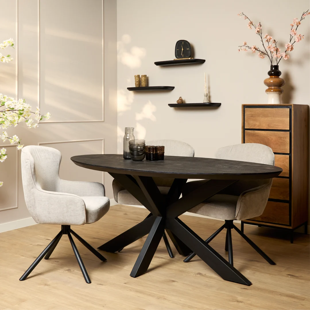 Een moderne eethoek met de Eettafel Brandy Zwart ovale tafel (180 cm), drie lichtbeige draaistoelen, een houten ladekast, wandplanken met decor en een vaas met takken, alles op een lichthouten vloer.