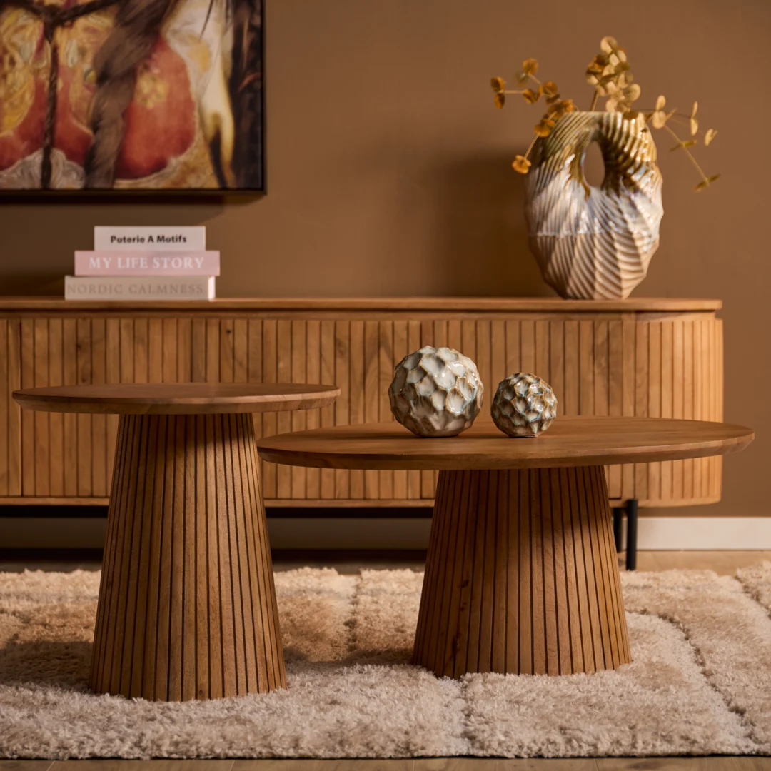 Twee houten salontafels met een rond blad en een geribbelde voet. Decoratieve objecten en een vaas op de tafel. Zachte belichting, warme kleuren.