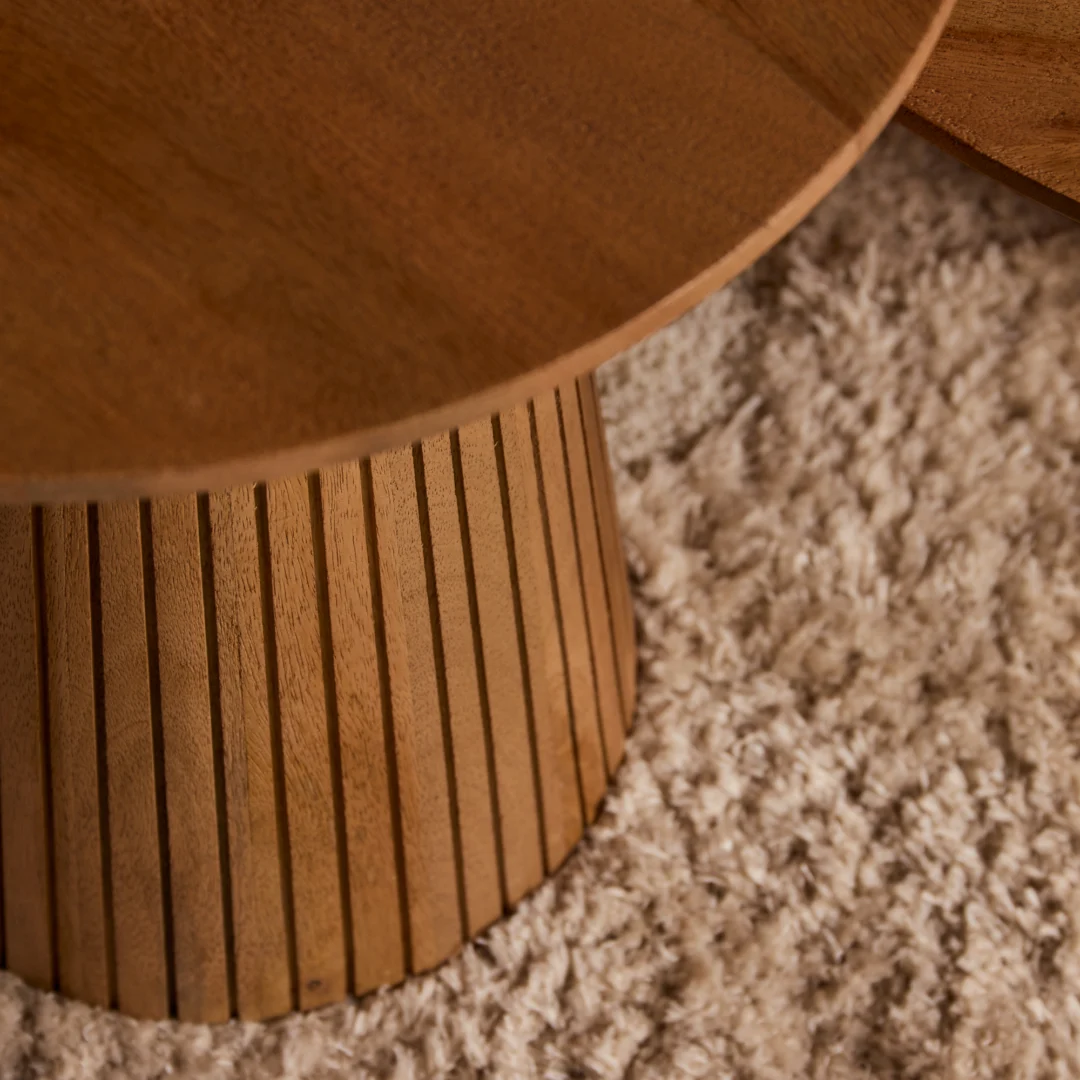 Detailopname van een ronde salontafel van hout met een roestkleurige afwerking, geplaatst op een zachte, crèmekleurige vloerkleed.