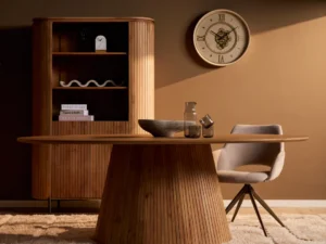 Ovale eettafel Solana van natuurlijk, robuust hout met verticale latten. Zachte belichting, beige stoel en kleed, bruine achtergrond met klok.