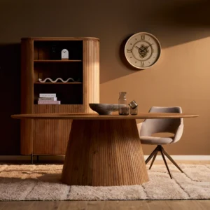 Ovale eettafel Solana van natuurlijk, robuust hout met verticale latten. Zachte belichting, beige stoel en kleed, bruine achtergrond met klok.