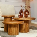 Drie Salontafel 40 CM ronde houten salontafels van verschillende hoogtes staan gegroepeerd in een woonkamer. Twee bruine keramieken vazen met rode bessen rusten op de hoogste tafel, met een beige bank op de achtergrond.